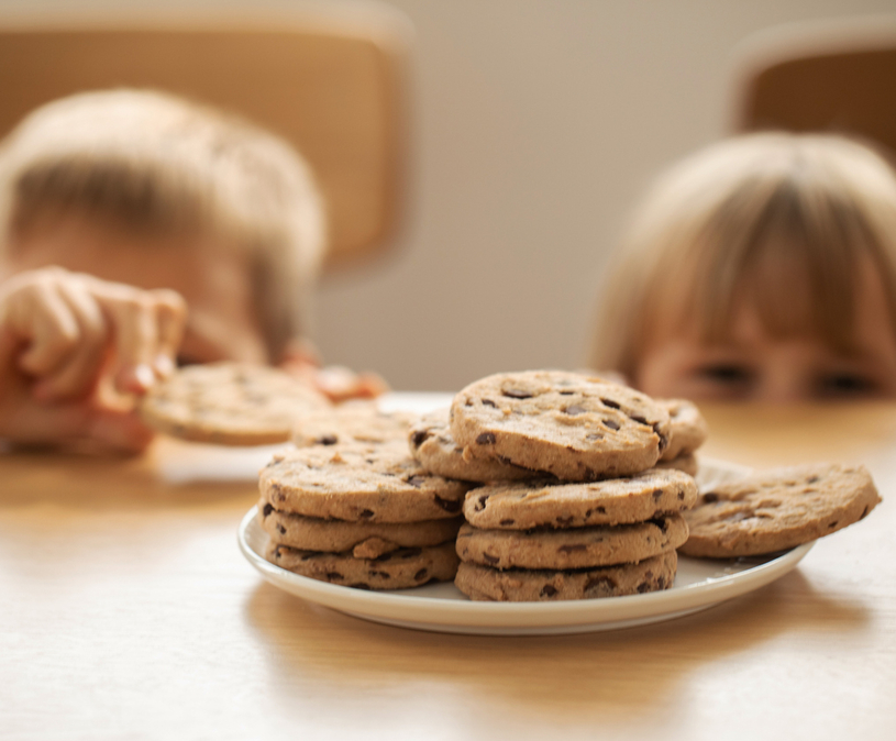 2 kinderen met blond haar steken uit boven een tafel waar een bord met koekjes opstaat